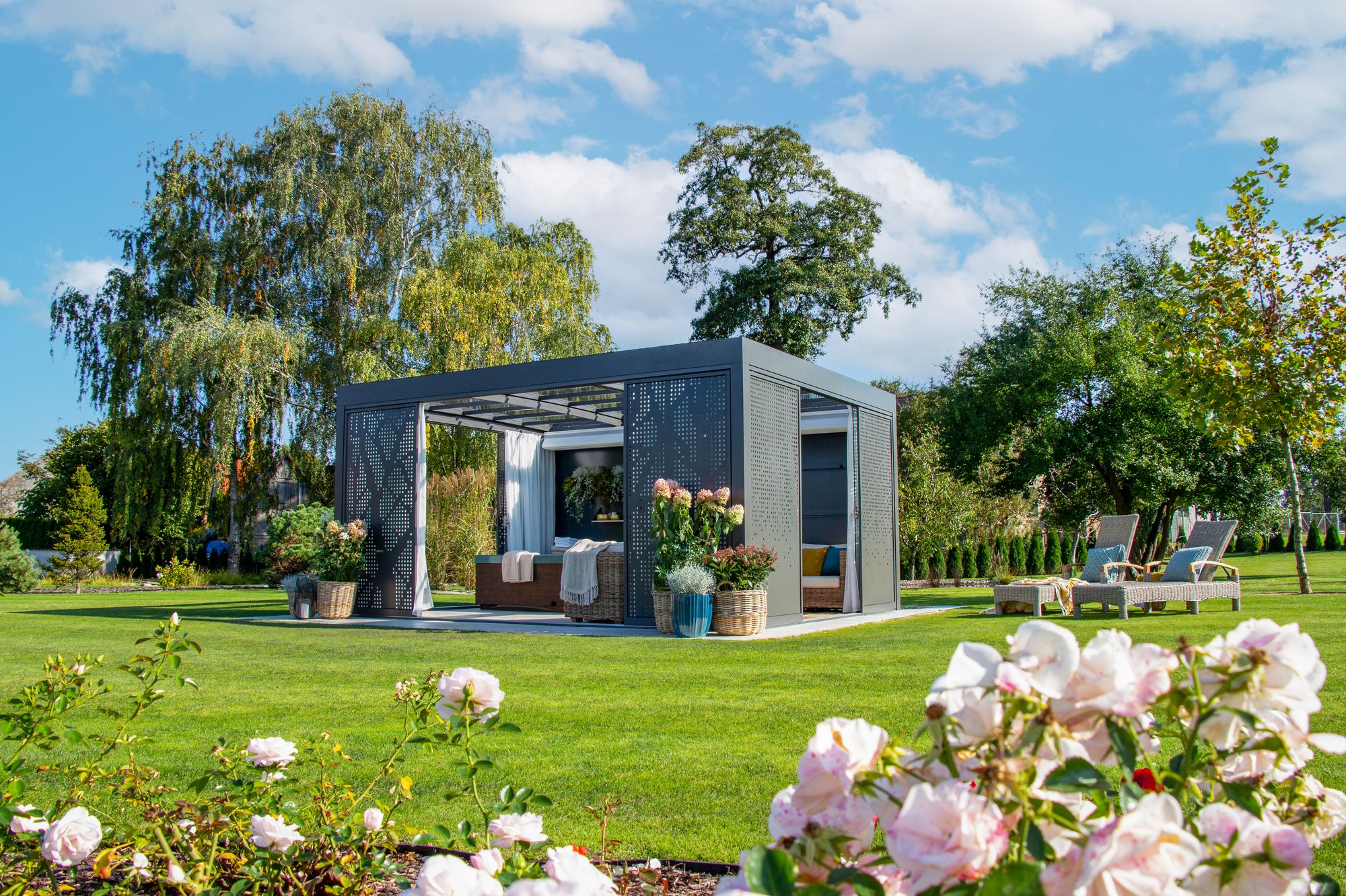 A freestanding glass room on a lawn, trees behind it and roses in the foreground