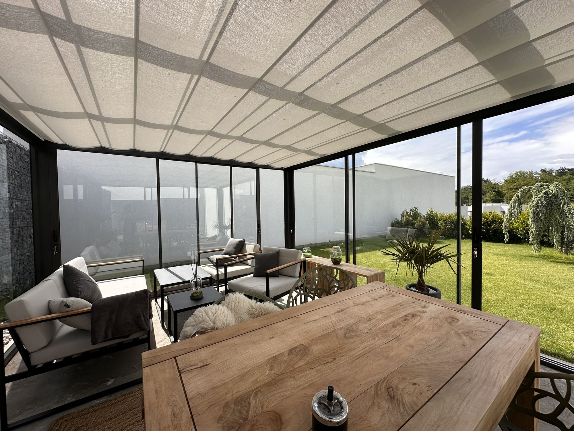 Inside a glass room with the fabric blinds drawn under the roof, a timber table in the shade